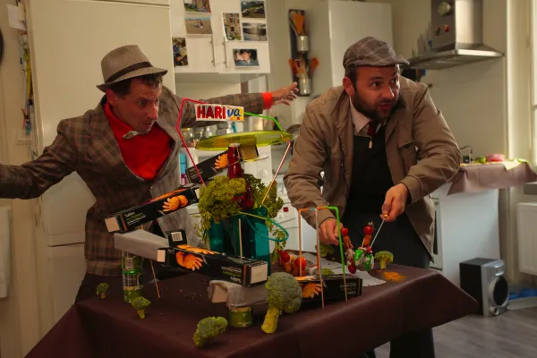 Photo  de deux comédiens autour d'une structure  construite en paquets de gâteaux, pailles et autres objets alimentaires. 