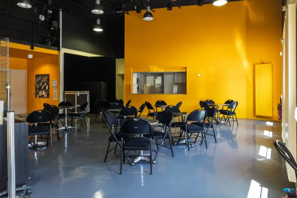 Photo d'une salle avec des tables rondes et des chaises, et un espace buvette au fond. 